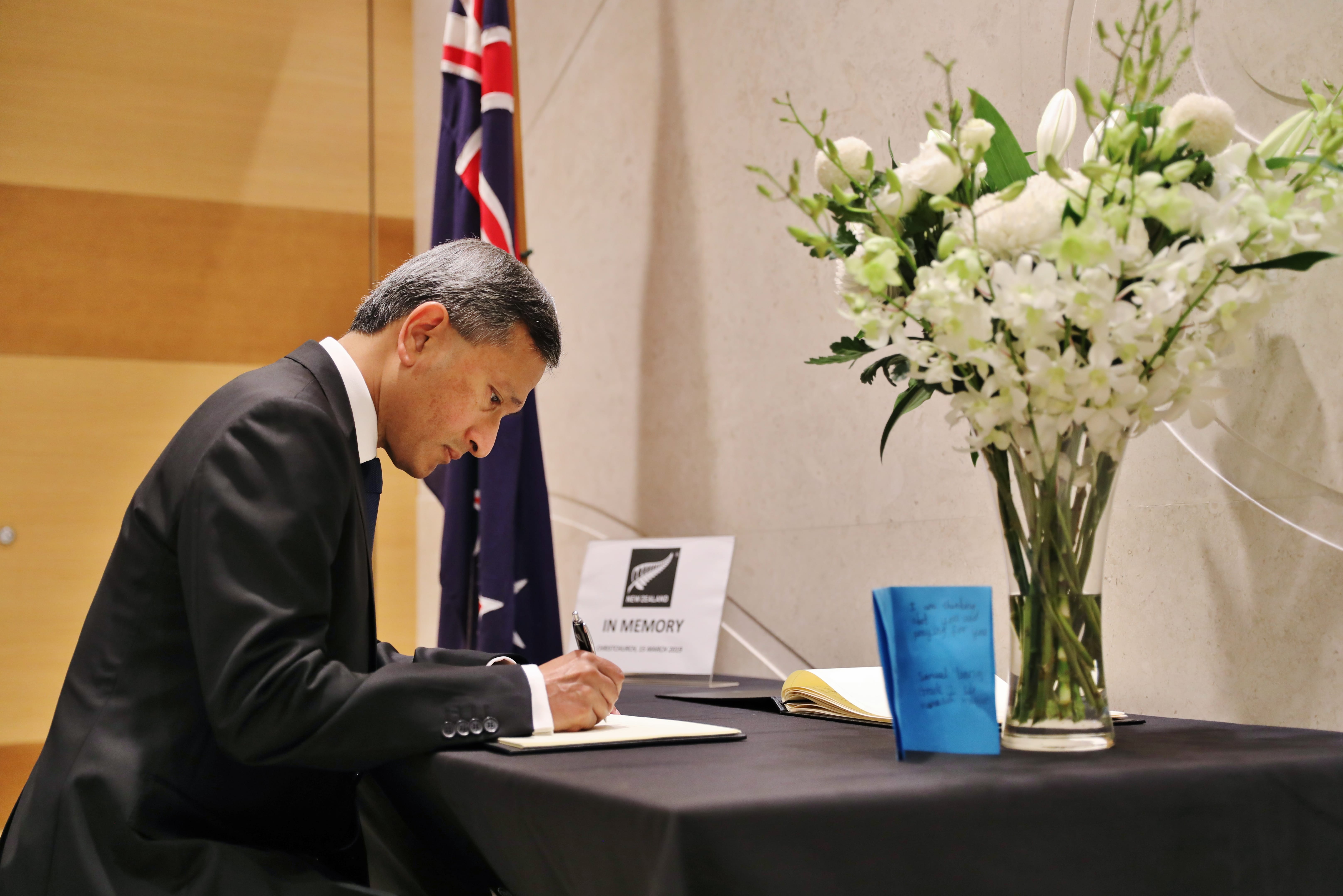 Man in suit signing guestbook. New Zealand flag, white flowers, "In Memory" sign on table.